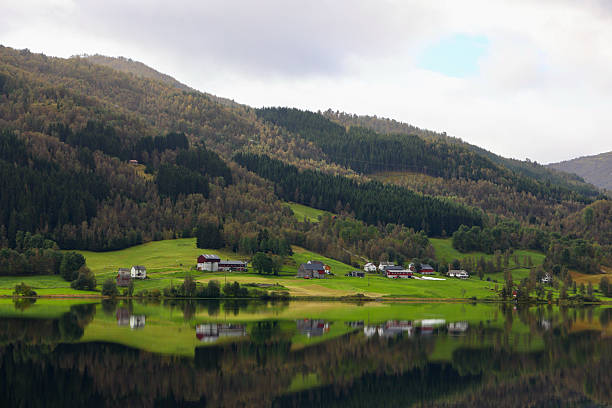 A serene landscape showcasing the peaceful shoreline of Oppheimsvatnet in Voss, Vestland county, Norway. Traditional farmhouses and barns rest on rolling green fields at the base of densely forested hillsides, perfectly mirrored in the still, glass-like lake. The soft light and muted clouds create a tranquil, atmospheric scene that highlights the untouched beauty of rural Western Norway. Ideal for themes of nature, countryside living, tranquility, and Scandinavian landscapes.
