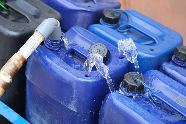 istock Hand-held hose fills multiple blue plastic jerry cans with water, causing some to overflow, indicating storage or distribution activity. 2247101134