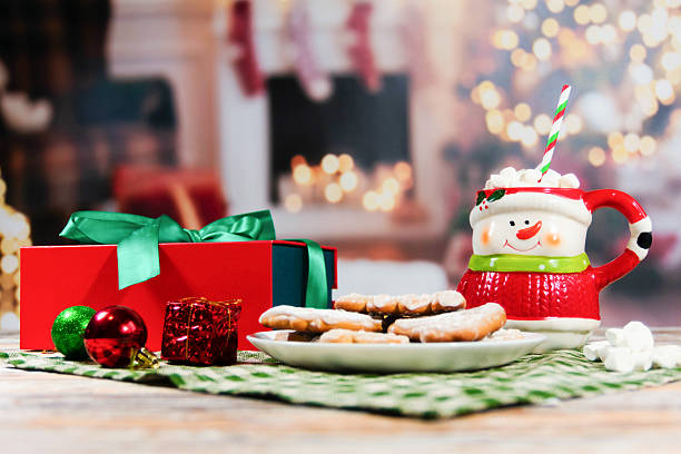 Plate of powdered cookies, snowman mug with marshmallows, gift boxes, and ornaments on wooden surface, festive indoor setup with blurred fireplace and Christmas tree in background.