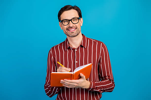istock Young handsome man with striped shirt writing in orange notebook against blue background 2247102586