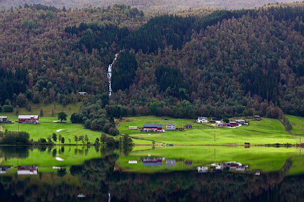 A peaceful rural landscape overlooking Oppheimsvatnet in Voss, Vestland county, Norway. Lush green fields, traditional farm buildings, and scattered houses stretch across the shoreline, while a narrow waterfall cascades down the densely forested hillside. The still lake surface creates a vivid mirror reflection of the farms, trees, and mountains, enhancing the calm, scenic atmosphere. This image captures the quiet beauty of Norwegian countryside life, natural harmony, and untouched Nordic scenery. Ideal for themes of nature, rural living, travel, and environmental serenity.