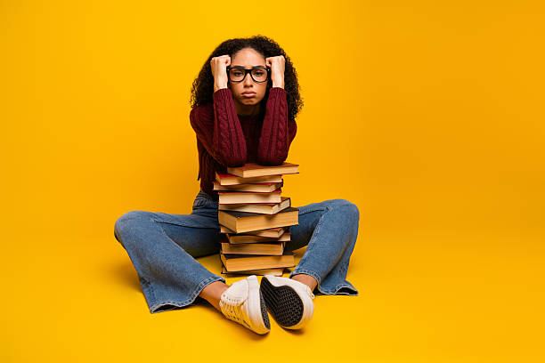 istock Young female student with glasses sits on the floor leaning on a tall stack of book against a bright yellow background 2247103867
