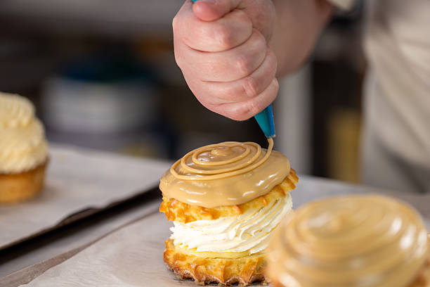 Close-up shot of cream cakes on a tray in a Northumberland bakery. There is an unrecognisable chef using a piping bag to decorate the cake with icing, ready to be sent to a local café.Videos similar to this scenario available.
