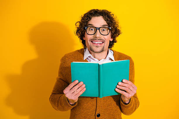 istock Young man with glasses reads turquoise notebook against a bright yellow background smiling for stock photography 2247107858