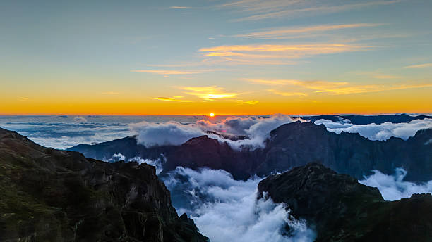 A dramatic orange sunset illuminates the sky above Pico do Arieiro in Madeira, with rugged mountains partially covered by clouds. Scenic landscape, atmospheric mountain view, natural beauty, travel destination