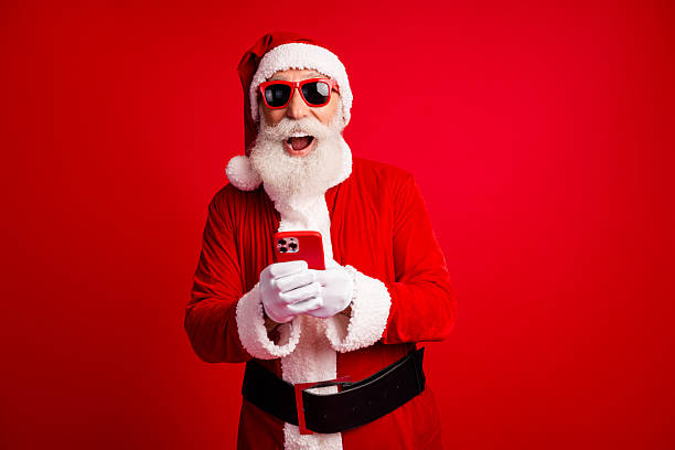 istock Santa Claus using a smartphone with cheerful expression wearing festive attire and sunglass on a red background 2247108123