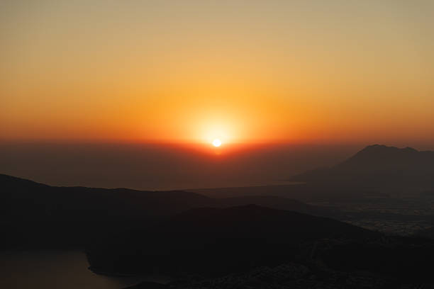 A tranquil sunset paints the sky in orange and gold as the sun sinks behind distant mountains. Dark silhouettes cradle a peaceful landscape, perfect for nature, travel, and coastal scenery projects