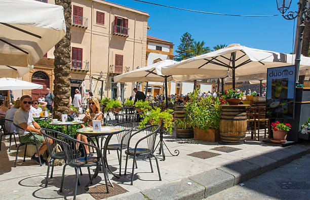 Cefalu, Italy - May 19 2025. Outdoor classic cafe-style bar seating in Piazza del Duomo, buzzing with tourists & locals enjoying food & drink under the shade of umbrellas. The architecture of the piazza provides a stunning historical context, with a 18th-19th century building with stucco facade, louvered shutters & balconies. A snapshot of daily spring life in Cefalu, where the grandeur of history provides a stage for a sunny afternoon
