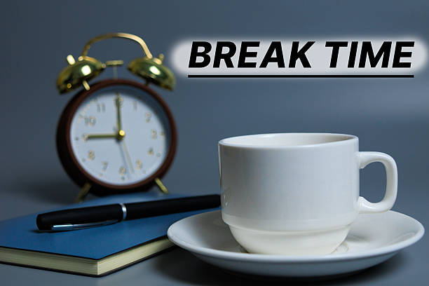 istock White coffee cup on a saucer beside a notebook and pen, with a blurred alarm clock in the background and the text “BREAK TIME,” 2247124099