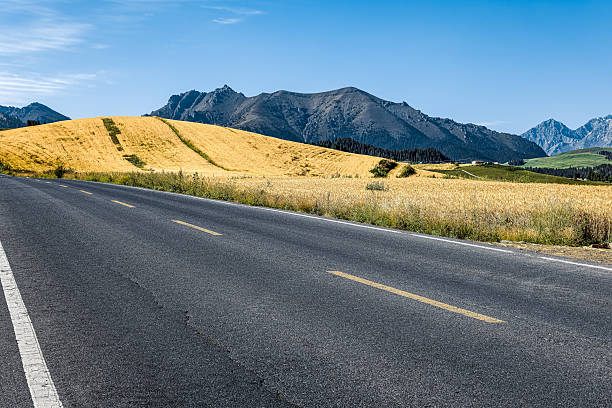 Paved road runs parallel to a golden hillside, with a stunning mountain range visible in the background on a bright day.