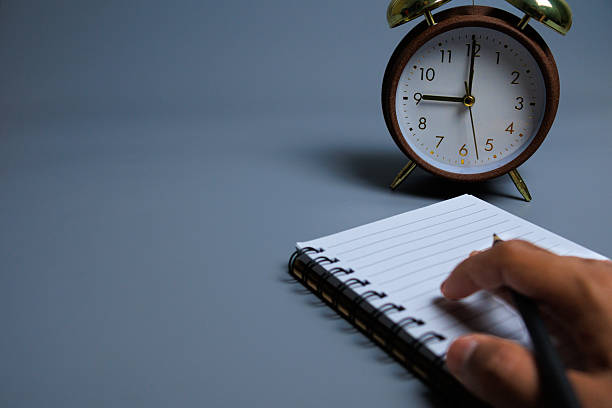 istock Hand writing with a pencil on a spiral notebook beside an alarm clock 2247124981