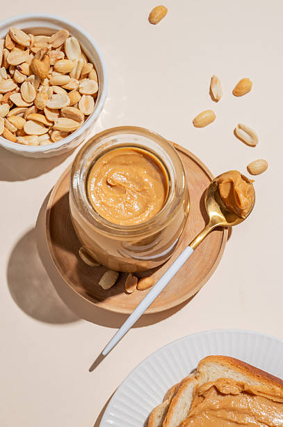 istock Peanut butter in a glass jar on a light background with scattered nuts, spoon and toast. 2247128196
