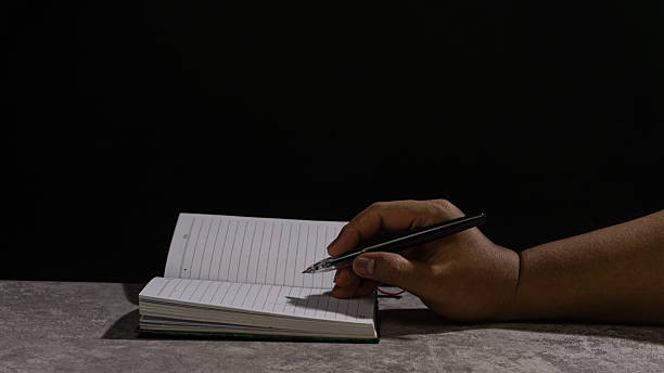 istock Hand holding a pen above an open notebook, lit dramatically against a dark background. Emphasizes anticipation, planning, and the start of a writing task. 2247130984