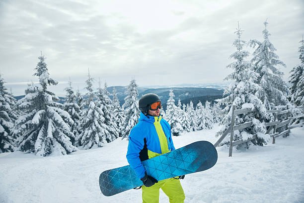 A snowboarder wearing a bright blue and green outfit is standing on a snowy slope holding a snowboard. Tall snow-covered trees surround the area.