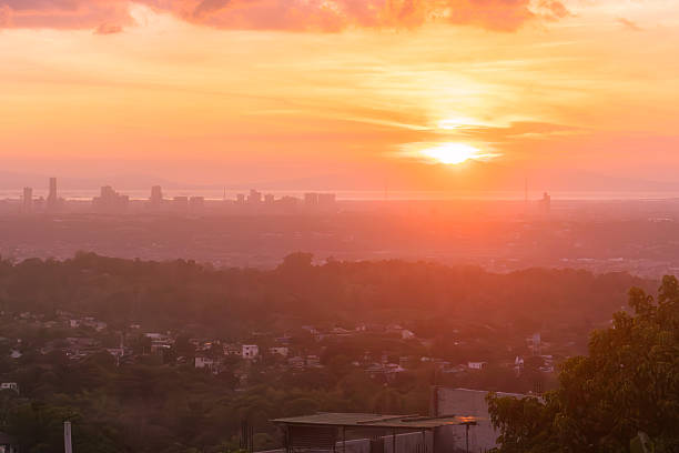 A soft, hazy sunset casts a warm orange and pink glow over a sprawling urban cityscape. The distant skyline and rolling hills are layered in the thick, atmospheric haze, creating a dreamy and serene view from a high overlook as the sun dips toward the horizon.