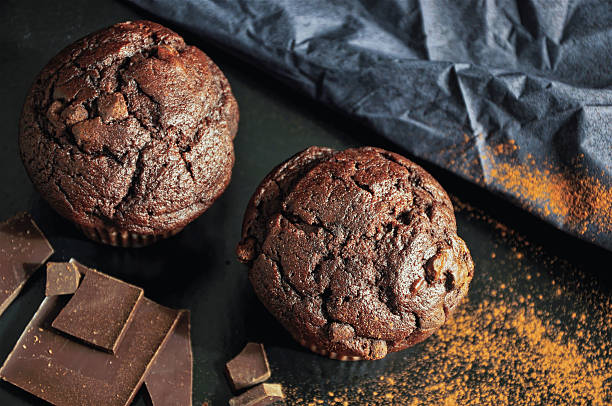 Top view of two decadent chocolate muffins surrounded by cocoa powder, chocolate chunks, and wrinkled cloth. A cozy and slightly rustic setup evoking handmade warmth and nostalgic baking. Copy space on upper and side areas.
