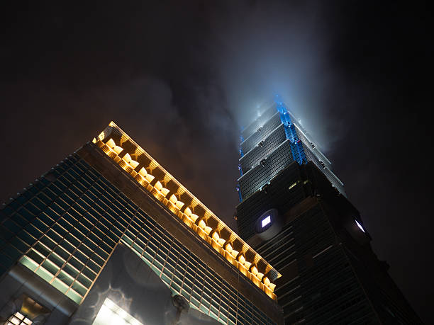 Taipei, Taiwan - November 16 2024: A dramatic low-angle view of Taipei 101, with the adjacent mall's lights and the tower vanishing into fog.