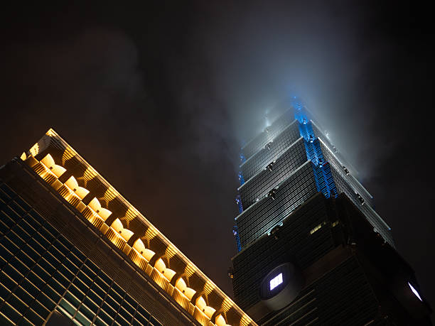 Taipei, Taiwan - November 16 2024: A dramatic, low-angle shot of Taipei 101, with the illuminated mall roof and the tower disappearing into clouds.