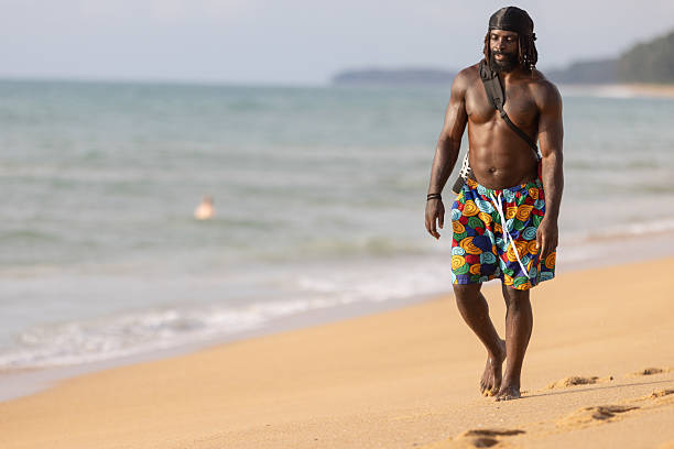 A smiling Black male in casual beachwear revels in the shallow sea water, radiating happiness during seaside leisure at a tropical beach.