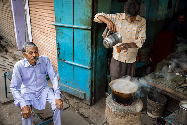 A street-side tea vendor pours steaming chai from a metal kettle into a small clay cup above a blackened pot set on a makeshift stove. Wisps of steam rise into the air while a man in a pale kurta-pajama sits nearby on a low metal stool, watching. Weathered blue doors, shutters and scattered utensils frame the compact, colorful stall, conveying a quiet moment of everyday street life.