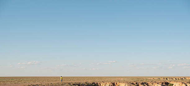 The vast expanse of the Outback stretches along the iconic 90 Mile Straight, revealing a dry and rugged terrain dotted with low shrubs and sparse trees under a clear blue sky.
