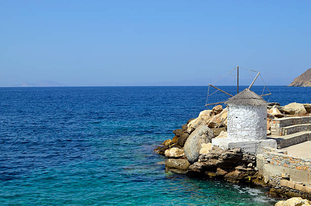 Amorgos, Greece - View of a small windmill at the jetty in the port of Aigiali with the clear blue Aegean Sea in the background.