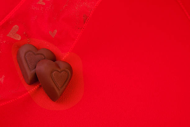 Two heart-shaped milk chocolate on red sheer ribbon with hearts on red fabric background. Studio shot with copy space.