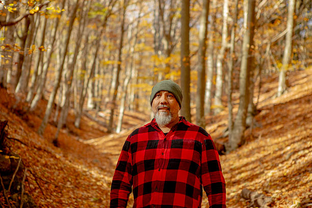 A man in a red plaid shirt walks along a leaf-strewn trail in a sun-dappled autumn forest, surrounded by trees with golden foliage.