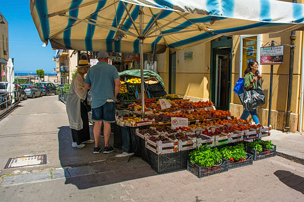 Cefalu, Italy - May 19 2025. A small sidewalk fruit & vegetable stall in the historic seaside town of Cefalu in early summer. A vibrant display of fresh seasonal Sicilian produce, it is laden with crates of colourful fruits, including various types of tomatoes, peaches, lemons & cherries. Pots of fresh basil, a staple of Italian cuisine, are also offered for sale. A couple of tourists browse the selection. An authentic & everyday scene