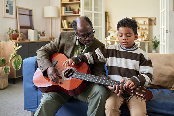 African American schoolgirl with acoustic guitar looking at paper with musical notes while playing instrument against classmates
