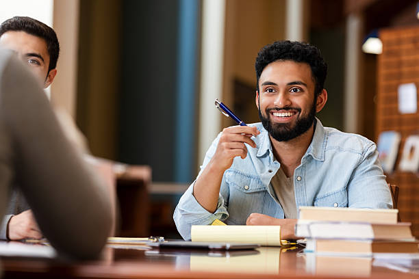 istock Smiling young man with beard holding a blue pen, discussing ideas with colleagues in library 2247174144