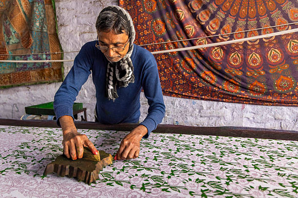 An artisan carefully hand-block prints a floral pattern onto a white fabric, pressing a carved wooden block with green dye across a long worktable. He wears glasses, a patterned scarf and a blue sweater, concentrating on aligning the design while colorful printed textiles hang on the workshop wall behind him.