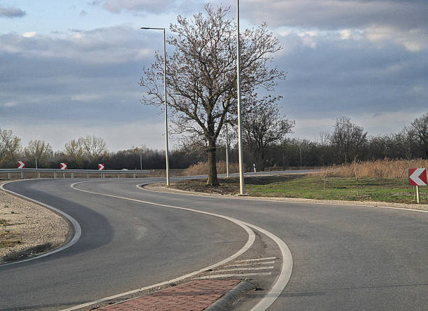 An empty S-shaped asphalt road winds sharply through a rural landscape, marked by white boundary lines and red-and-white chevron directional signs under a gloomy overcast sky. This transportation scene illustrates concepts of driving safety, navigating dangerous turns, and rural infrastructure during the late autumn or winter season.