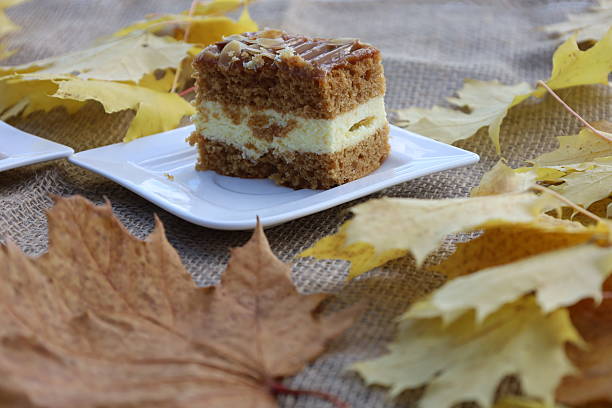 A layered cake slice with caramel glaze sits on a white plate on burlap. Yellow and brown dry autumn leaves scattered around create a cozy fall atmosphere.