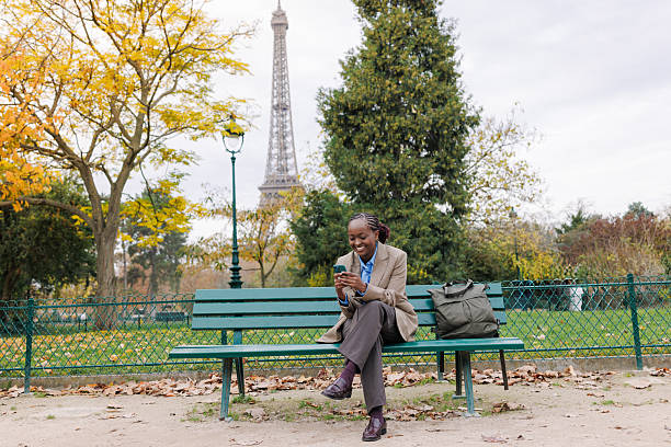 A young Black woman wearing a beige coat sits on a green park bench in Paris, using her smartphone. The autumn setting includes yellow leaves and the Eiffel Tower behind her