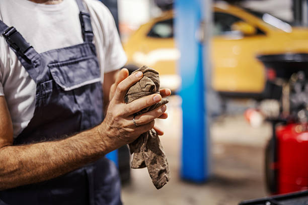 Mechanic wiping dirty hands with a rag in an automotive repair workshop. He is wearing blue overalls, cleaning up after car maintenance