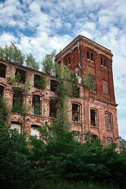 Abandoned red-brick industrial building with broken windows and vegetation overtaking the facade, illustrating architectural decay and historic character in Poland.