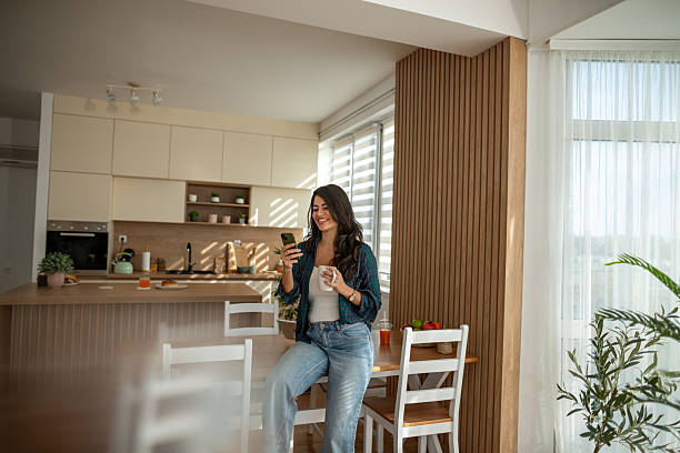 istock Young woman smiling, using smartphone and drinking coffee in kitchen 2247191799