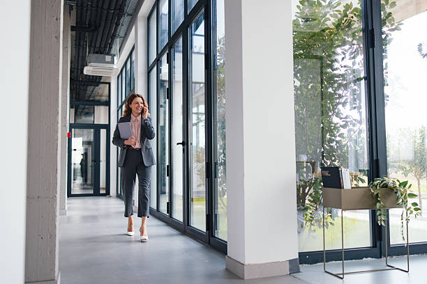 istock Professional Businesswoman Walking Through Modern Office Corridor On Phone With Folder And Glass Windows 2247197771