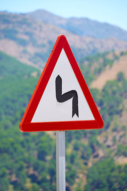 A prominent triangular red and white road sign warning of a sharp curve ahead, set against a vivid landscape of green hills and mountains with a bright blue sky under daylight.