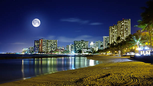 A captivating nighttime view of Waikiki Beach, located in Oahu, Hawaii, with a full moon casting reflections on the water and illuminated high-rise hotels creating a serene and picturesque tropical atmosphere.