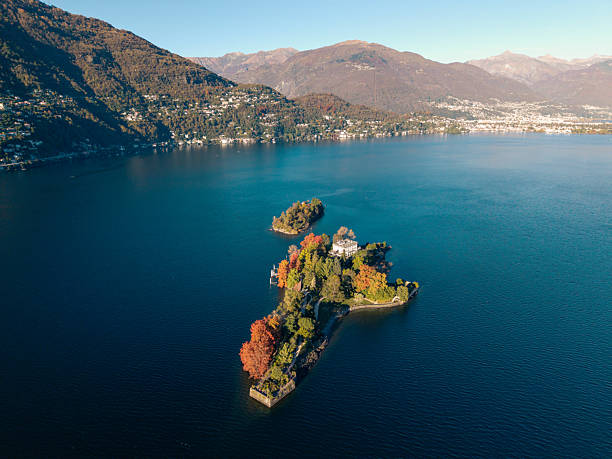 Aerial view of a small island covered with colorful autumn trees and a building surrounded by calm blue lake water. Scenic landscape with mountains and clear sky in the background. Nature and travel concept.