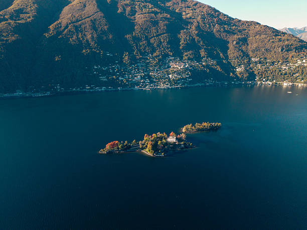 Aerial view of a small island with a villa surrounded by trees in a calm mountain lake. Scenic landscape with water, forested hills, and distant town on the shore. Nature and travel concept.