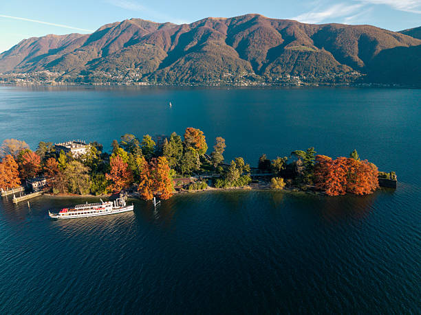Small island with colorful autumn trees and a docked boat on a clear mountain lake. Scenic lake view with mountains in the background. Peaceful nature landscape with water and forest.