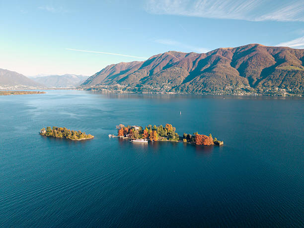 Two small islands with autumn trees in a calm blue lake surrounded by large mountains. Scenic aerial view of peaceful lake landscape with islands and distant mountain range. Beautiful nature and water scenery.
