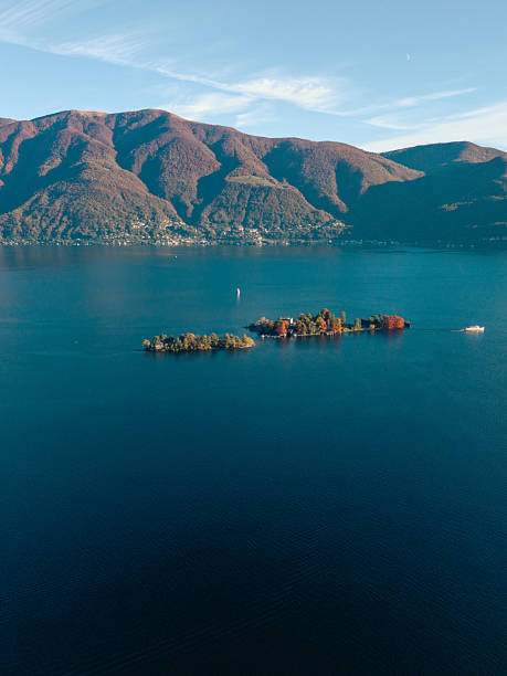 Aerial view of a small island with autumn trees in a calm blue mountain lake. Scenic landscape with boat approaching the island. Nature and travel concept with peaceful water and hills.