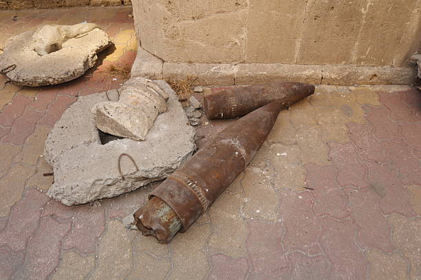 War remnants and unexploded artillery shells on a street in Aleppo, Syria, with civilians walking nearby — a powerful scene showing the danger and the urgent need for reconstruction and safety.