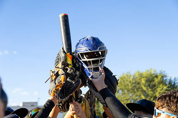 Baseball team hands lifting catcher's helmet, bat, and gloves together in celebration under a blue sky, symbolizing triumph