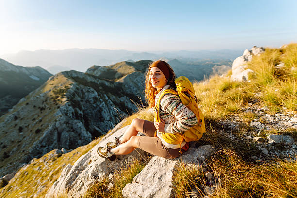 istock Beautiful female hiker with yellow backpack stands on mountain ridge, watching sunset. 2247248038