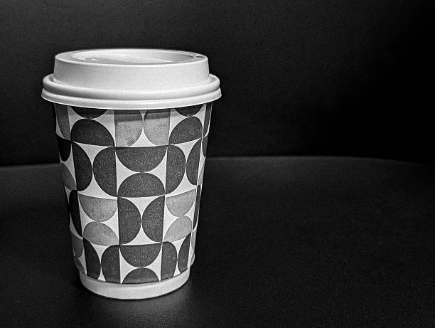 A sophisticated black and white (grayscale) photograph of a take-away coffee cup featuring a clean geometric pattern and a light-colored lid. The cup is centrally placed on a dark, plain background, offering generous negative space for text. This high-contrast, monochrome image is ideal for classic branding, formal editorial content, minimalist design, and advertising requiring a strong, uncluttered visual.